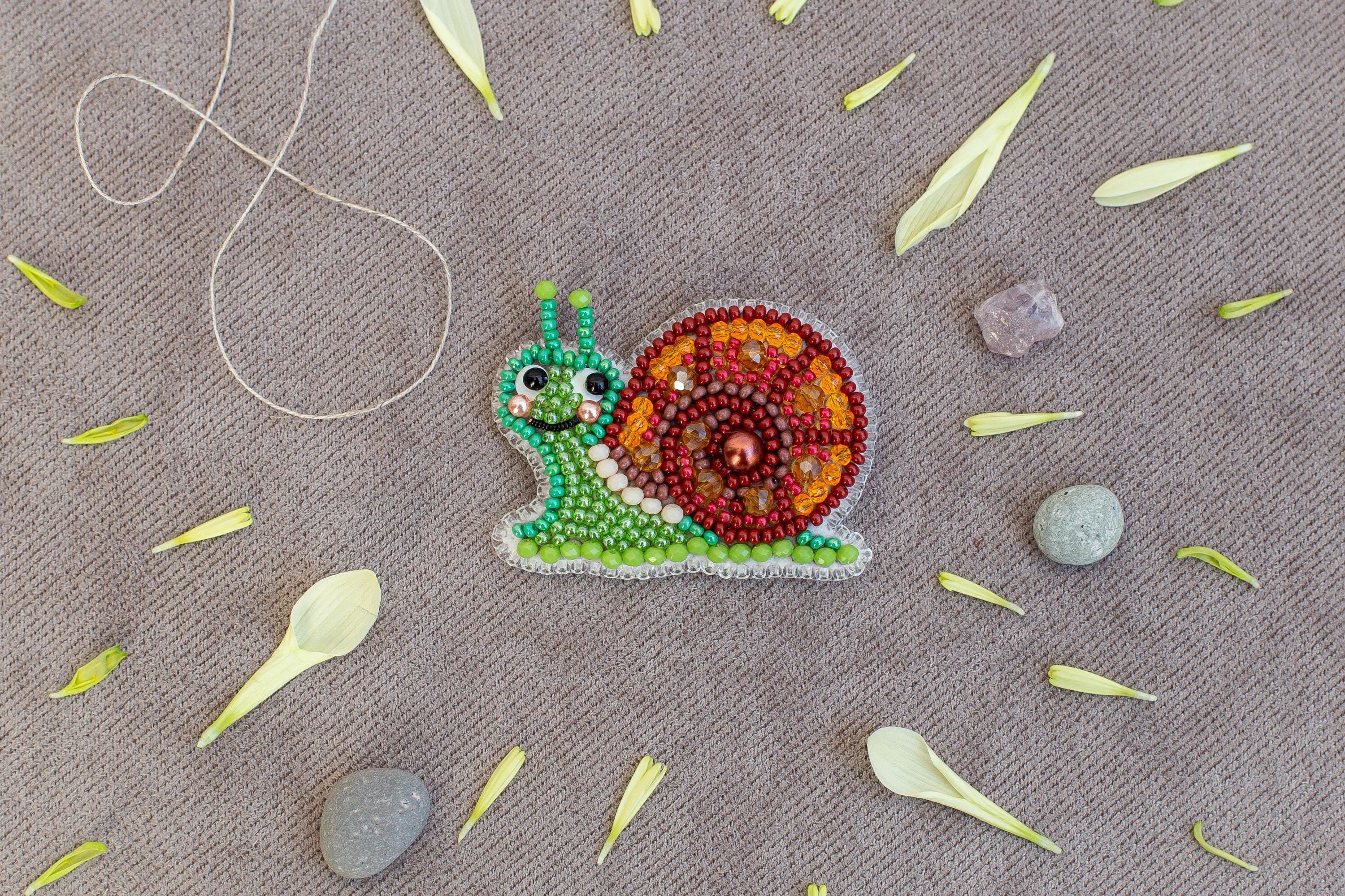 A colorful bead necklace depicting a snail is displayed on a beige surface, surrounded by scattered flower petals and small rocks.